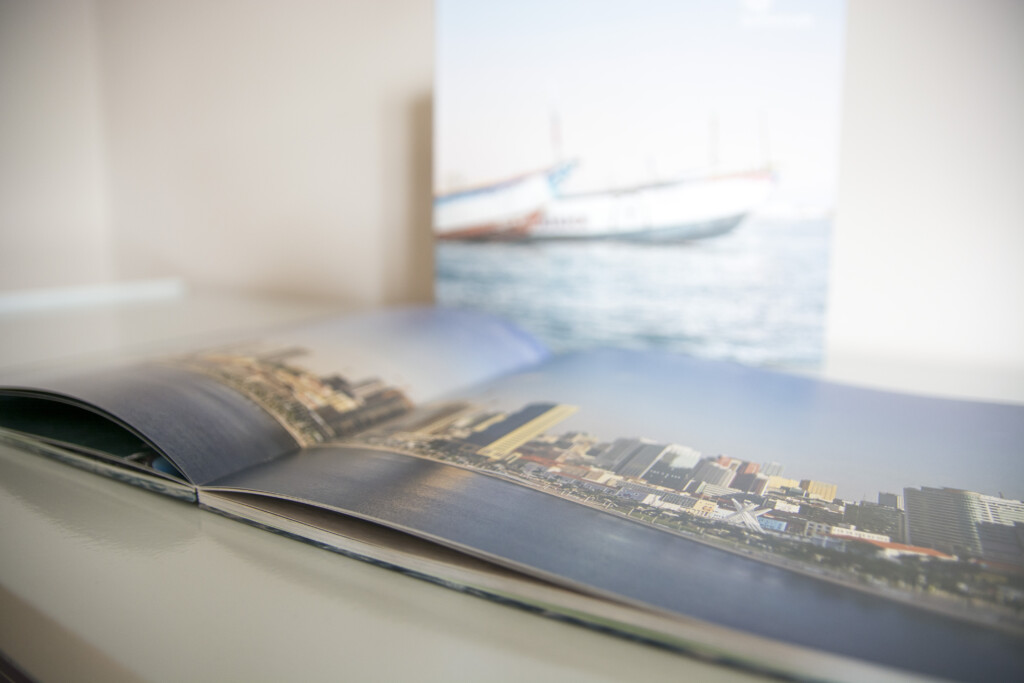 Open hardcover book with aerial cityscape photograph visible on spread, displayed on white surface with blurred seascape