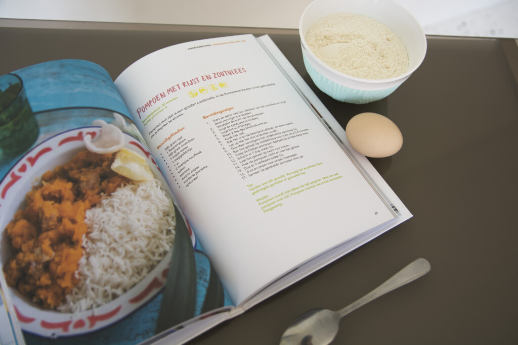 Hardcover cookbook open to a recipe page, displayed with a finished copy showing the cover and a bowl of prepared dish
