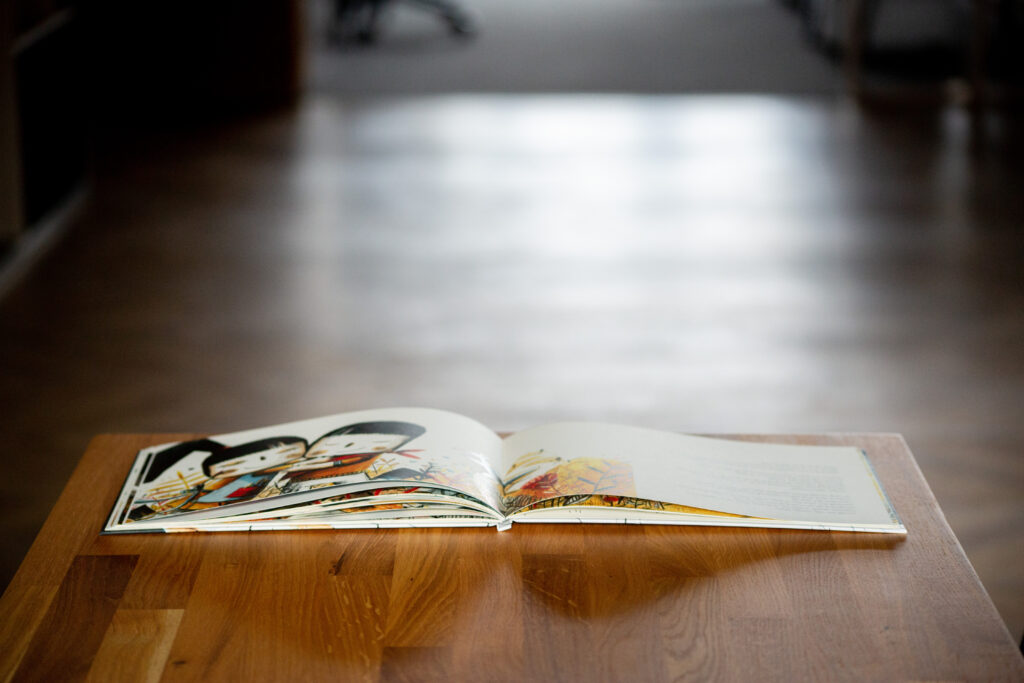 Open hardcover book with illustrated pages displayed on wooden table, interior pages visible in soft focus.