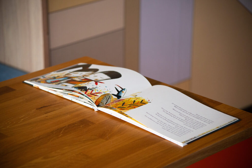 Hardcover children's book open on wooden table, showing illustrated pages with colourful artwork and text.