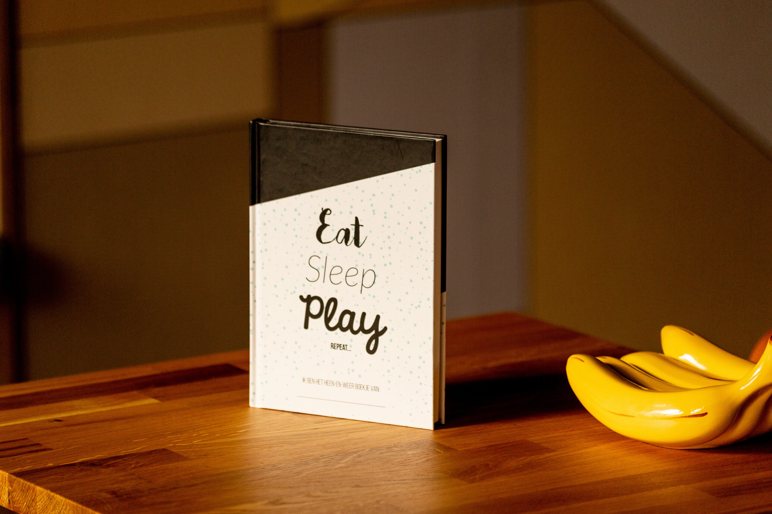 Hardcover book with black cloth spine and cream cover printed with "Eat Sleep Play" text, displayed on wooden table next to