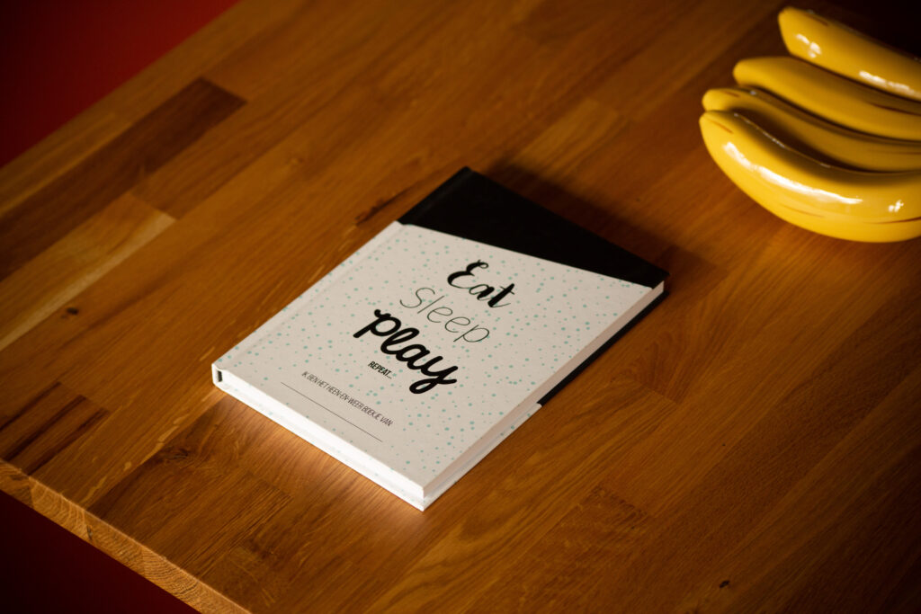 Hardcover book with white cover featuring black handwritten-style text reading "Eat Sleep Play" on wooden surface next to