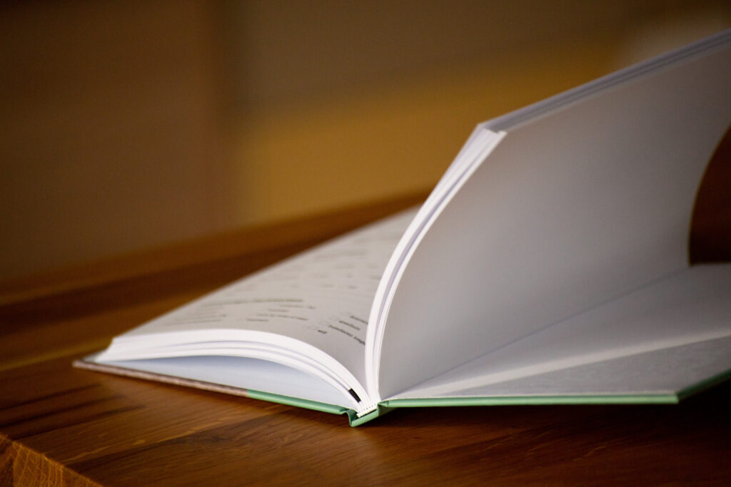 Open hardcover book with white pages on a wooden surface, showing binding and page edges.