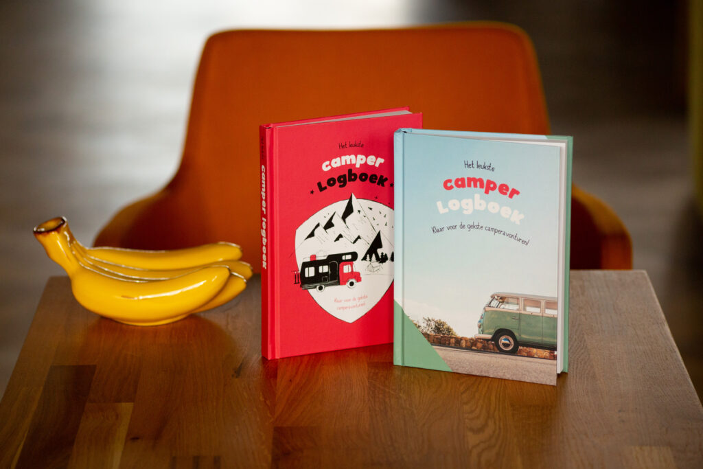 Two softcover books titled "camper logboek" in red and green, displayed next to a banana on a wooden table with an orange