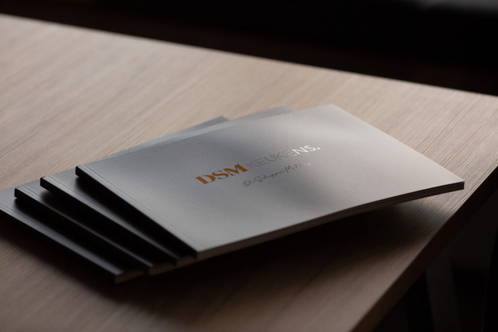 Stack of four printed catalogues with silver-grey covers and gold foil-stamped text, fanned slightly on a light wood surface.