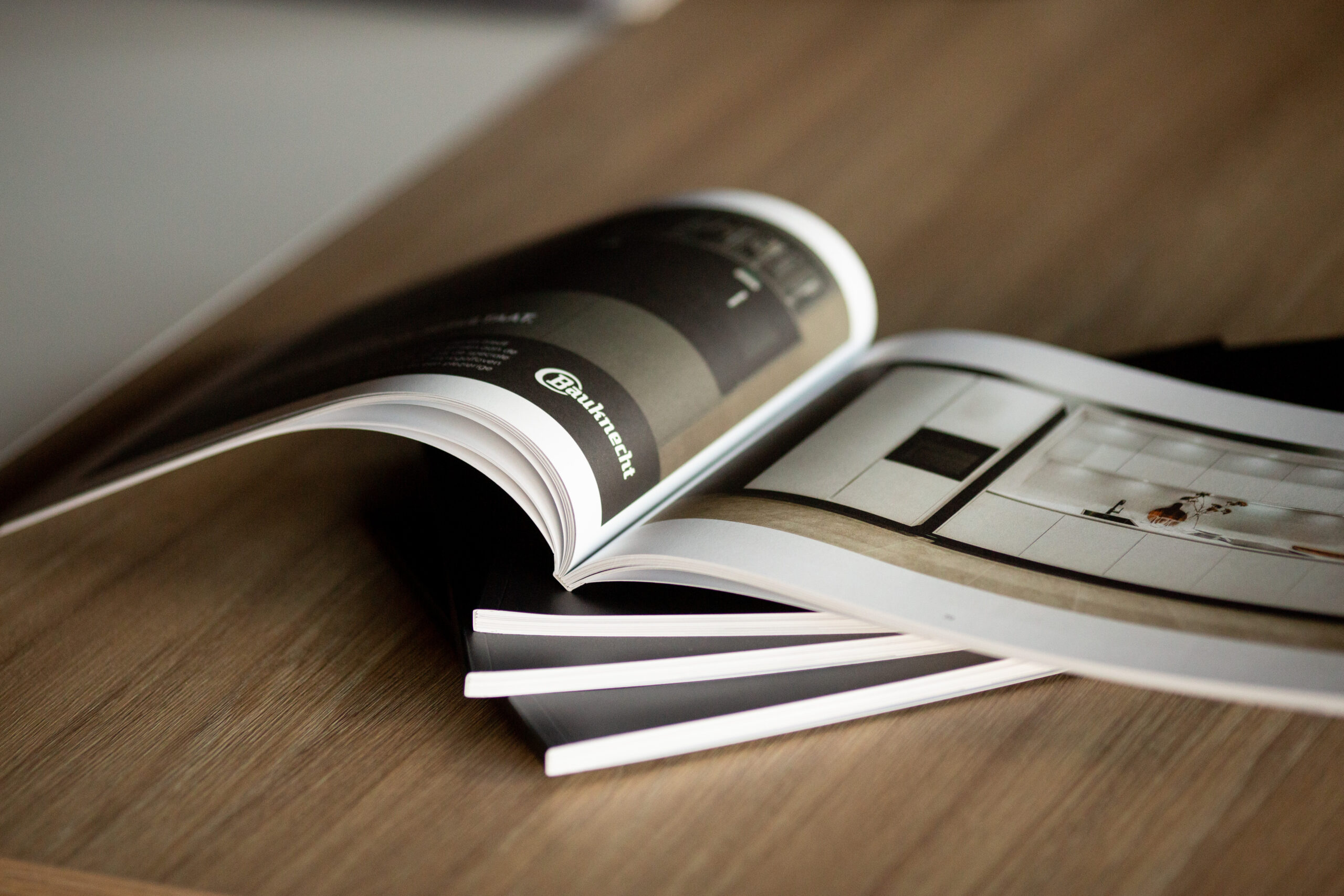 Open catalogue with gloss-laminated cover fanned beside stack of white pages on wooden surface.