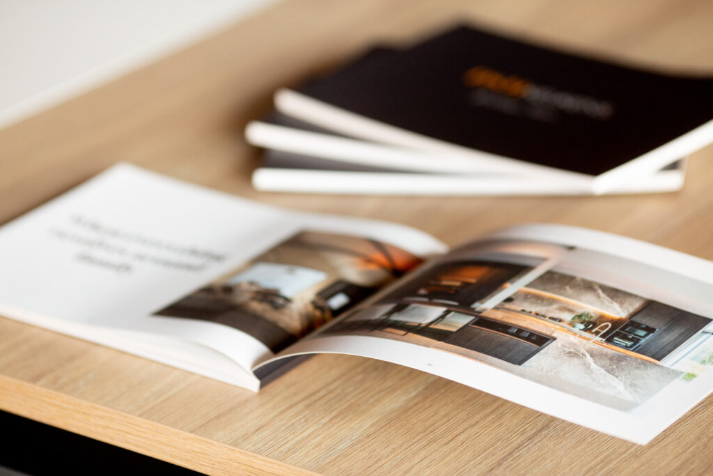 Printed catalogue with colour photography spread open on wooden table, showing interior pages with images of interiors and