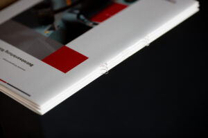 Printed brochure with white cover featuring red geometric shapes and detail of bound spine edge.