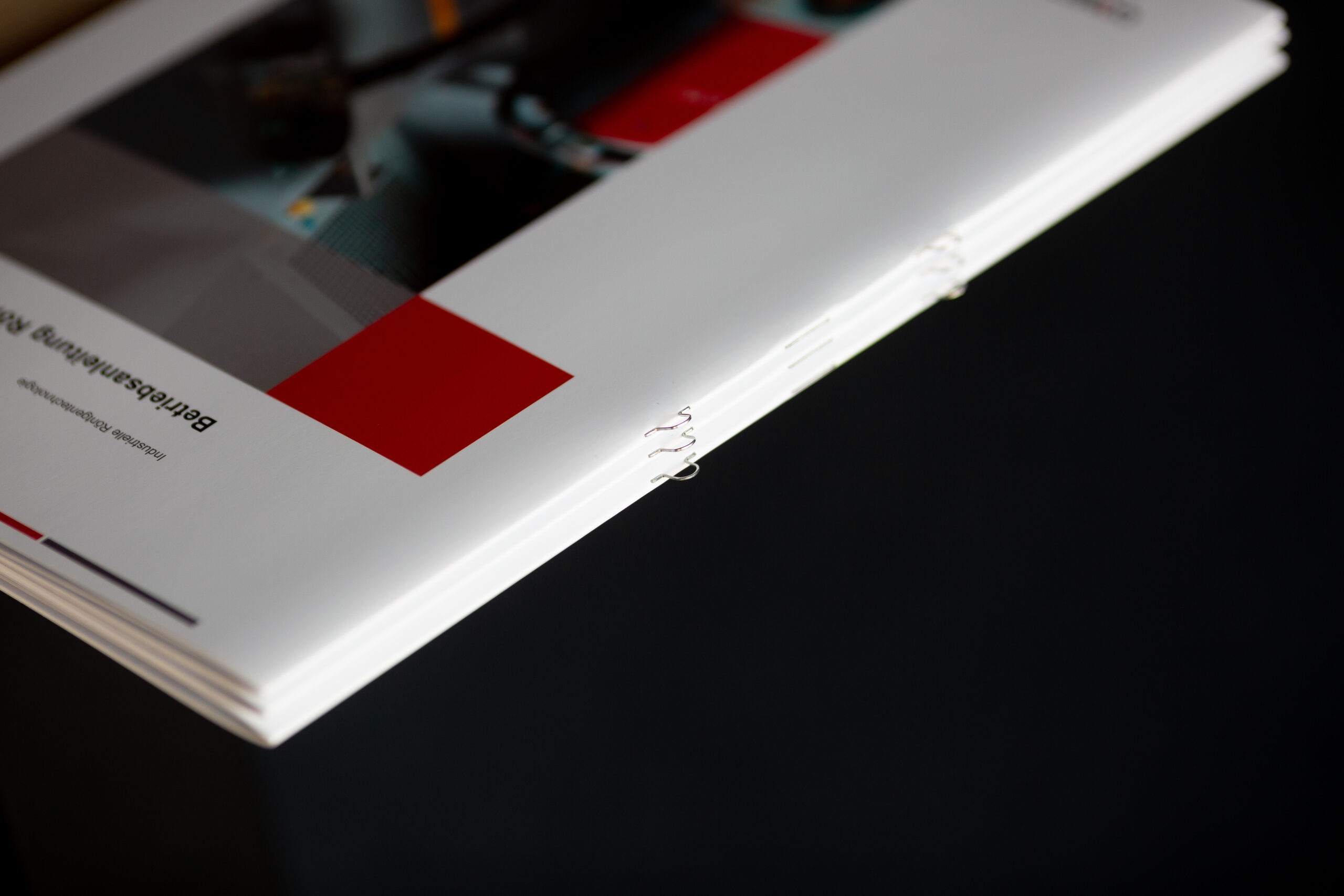 Printed brochure with white cover featuring red geometric shapes and detail of bound spine edge.