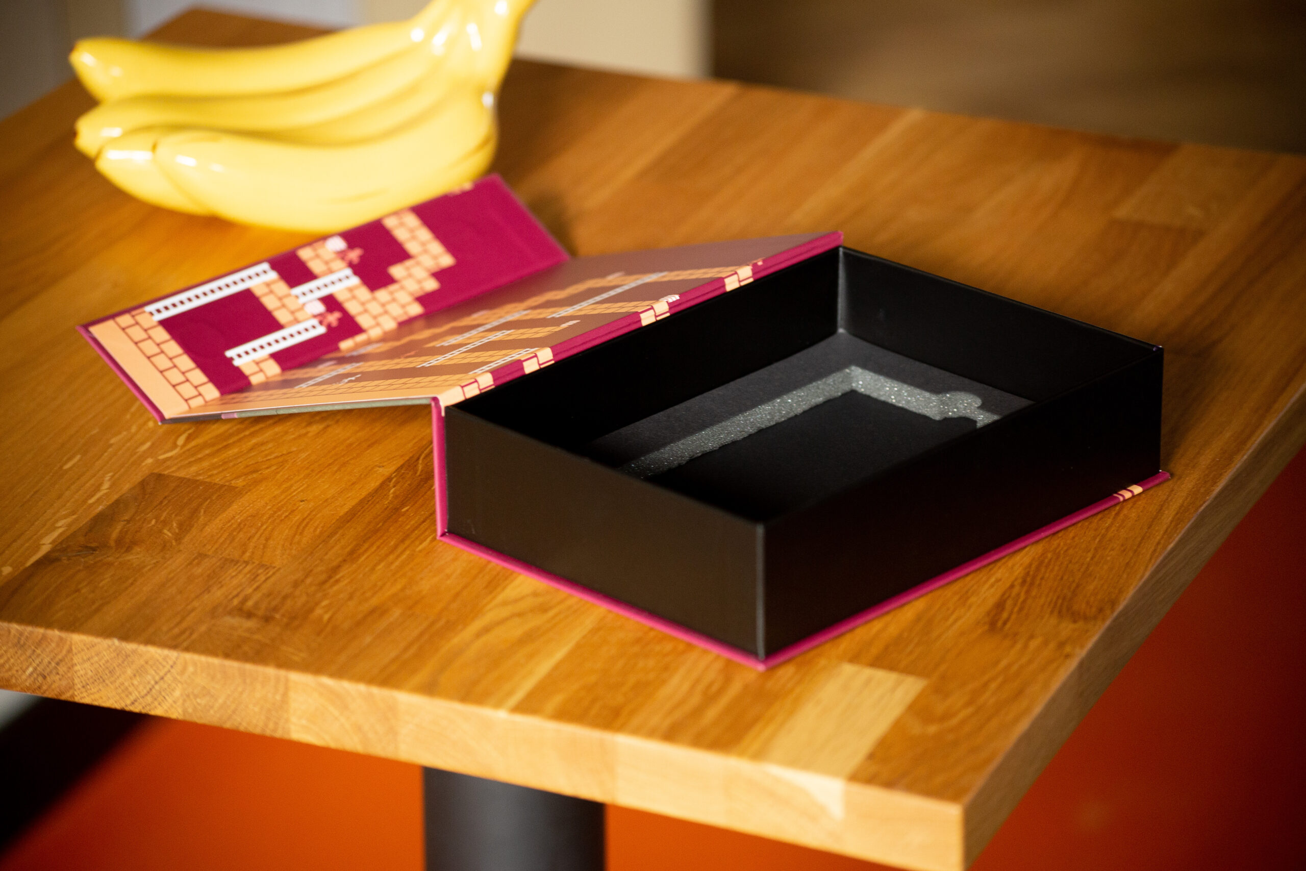 Black rigid box with pink interior and printed insert, shown open on wooden table with bananas and folded printed material