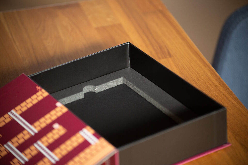 Open black box with grey interior padding on wooden surface next to burgundy hardcover books with gold embossed spines.
